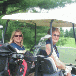 2 women sit in a golf cart, looking over their shoulders at the camera with big smiles