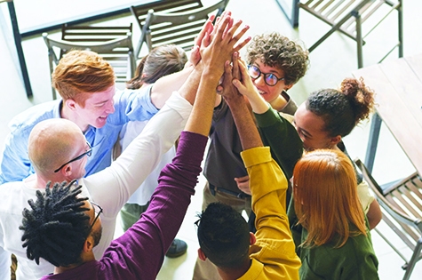 a group of 8 adults stand inside a bright lit room. They are each holding up a hand into the centre of the group all high-fiving eachother.