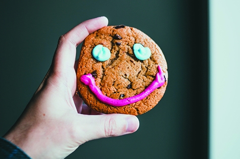 a hand coming from the bottom left of the frame holding a cookie. The cookie has a smile on it made from icing, two blue eyes and a pink smile.