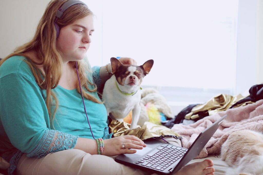 A woman sits inside on a bed with fluffy blankets and her relaxed dog. She is wearing a blue shit and beige pants and has a laptop on her legs with headphones on around her head. She is wearing a few bracelets and appears to have a wedding band on. Beside her is a loyal dog that she is petting and the dog has his eyes squinted closed and appears to be enjoying the pats.