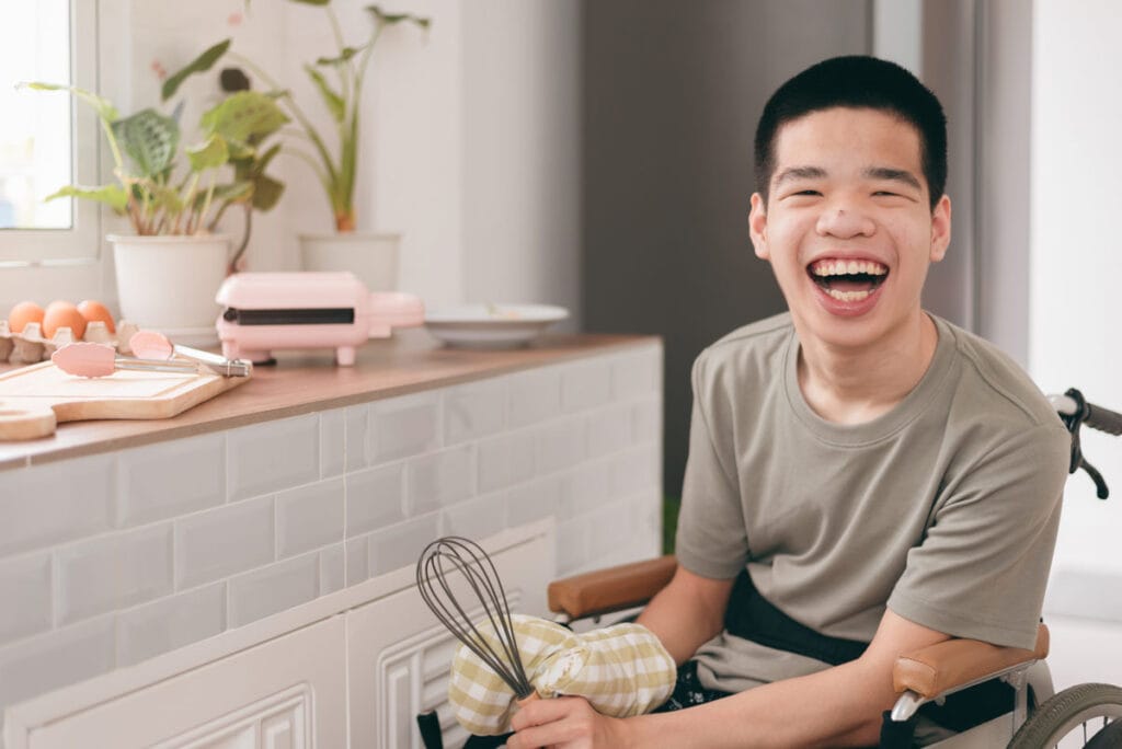 A man appears to be inside a kitchen. He is a young man with a huge smile on his face. He is sitting in a wheelchair wearing a grey tshirt. He has an oven mitt on one hand and is holding a whisk in his other hand. On the counter beside him there is a cutting board and tongs, a carton of eggs, a couple of plants and a sandwich press.