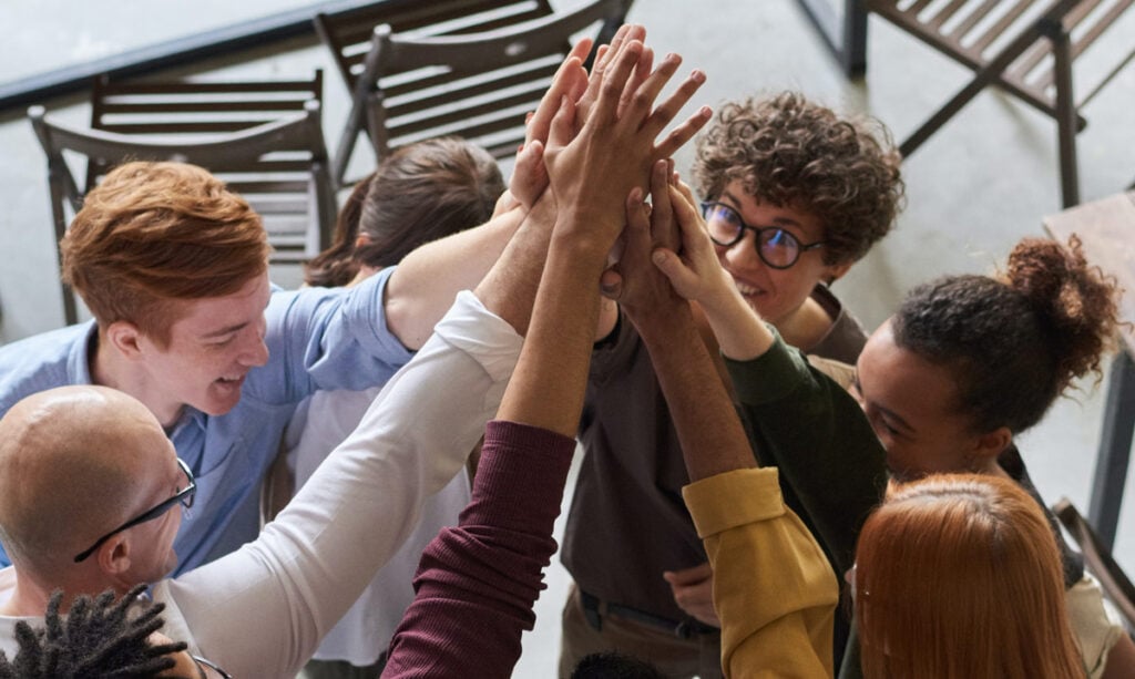 a group of people standing inside. In this angle we are looking down on them as they all high five together. The group ranges in diversity. There is a white man with red hair and a blue tshirt on the left. Next to him you can see the brown hair of a woman, her face is tucked down, and she is wearing a white tshirt. Next to her is an individual with brown curly hair atop their head. They are wearing round dark framed glasses. Next to them is a dark skinned woman with curly hair tied back in a pony tail. To the bottom right we see a white woman with red hair from behind. Next to her at the bottom left we can see a portion of a black man with his dark hair and dark framed glasses. And between this man and the first man with red hair, there is a bald gentleman with glasses.