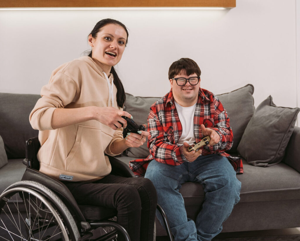 Two people sit together indoors playing video games. There is a young woman on a wheelchair wearing black jeans and a cream coloured sweater. She is holding a game controller in her hand and looking at the camera with a happy look on her face. The other person is sitting on a grey couch also with a game controller in their hand and also looking forward. They are wearing dark framed glasses and a red plaid shirt over top of a white tshirt and blue jeans.