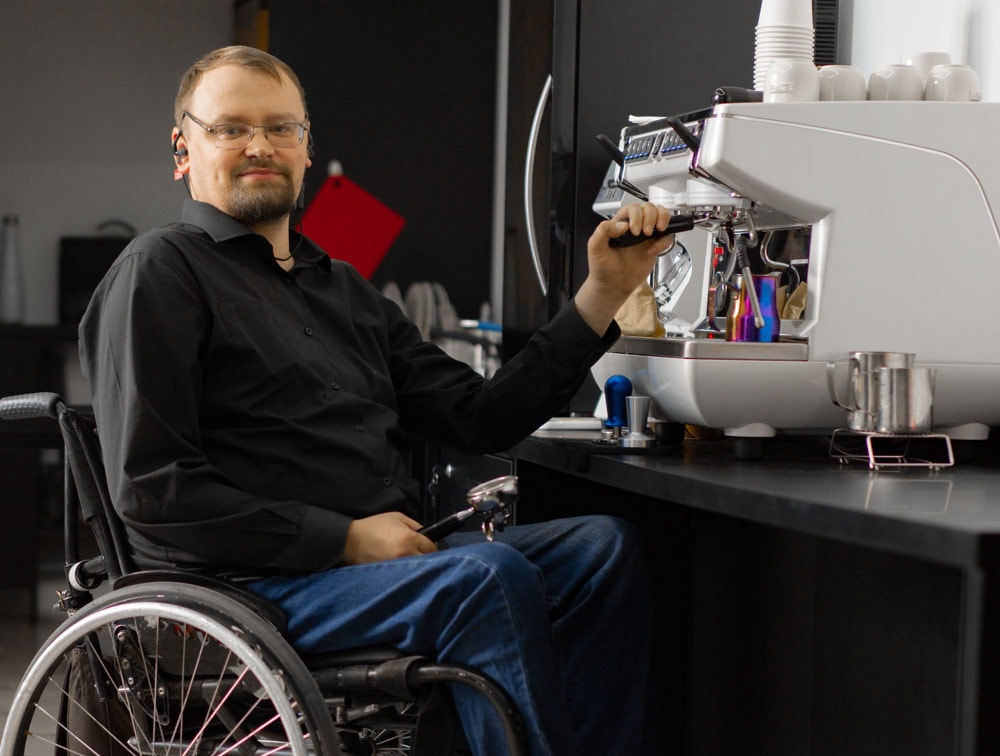 A man sits indoors making himself an espresso. He is sitting in a wheel chair and is wearing a black button down shirt and jeans. He has one hand in his lap holding onto some type of tool. His other hand is up on the espresso machine. He is wearing glasses and has facial hair.