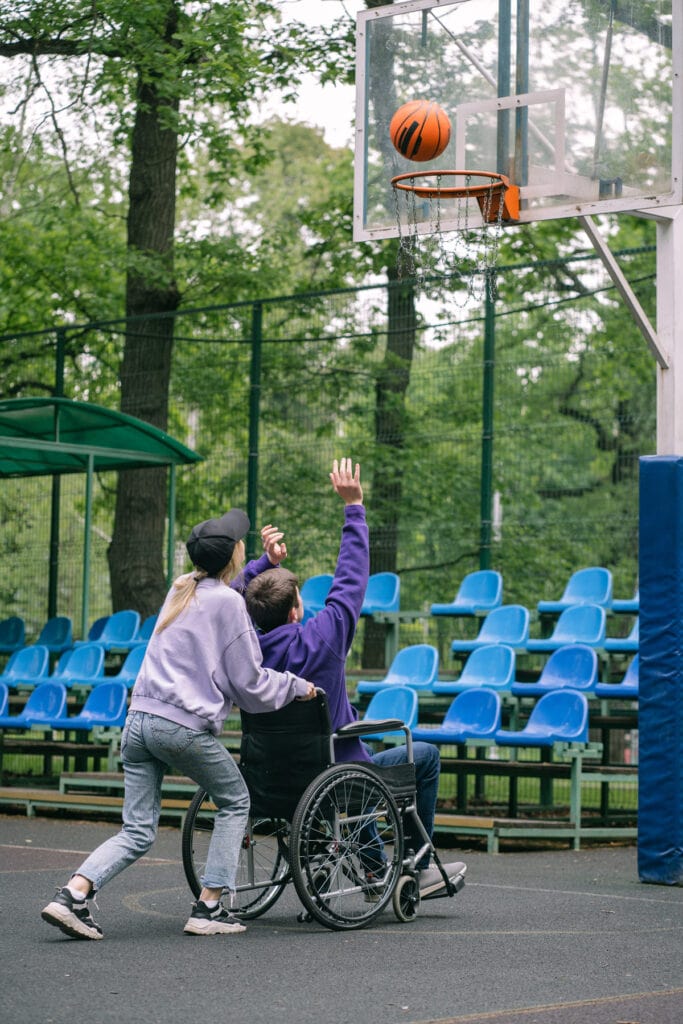 We see a woman from behind wearing a lilac coloured sweater, a black baseball cap and jeans and sneakers. She has long blonde hair tied back in a ponytail under her cap. She appears to be pushing a man in a wheelchair. The man is wearing a purple sweater and jeans and has his arms extended in the air as though he has just thrown a ball. There is a basketball in the air about to land in a basketball net. They are in a basketball court type area.