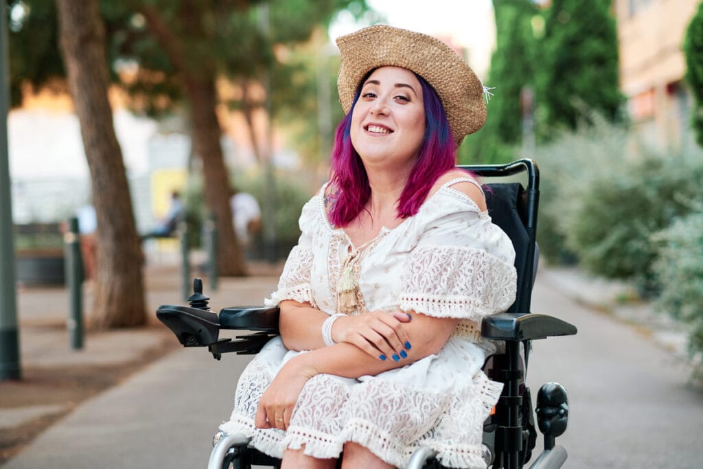 a woman sits comfortably in her wheelchair and smiles at the camera. She is sitting outside and you can see plants and trees blurred in the background behind her. She is wearing a rattan type hat and has beautiful bright purple and pink hair that is down past her shoulders. She is wearing a beautiful white lace dress that appears to fall just below her knees. She has one hand down on her leg and the other hand rests on her arm. She has bright blue nail polish on.