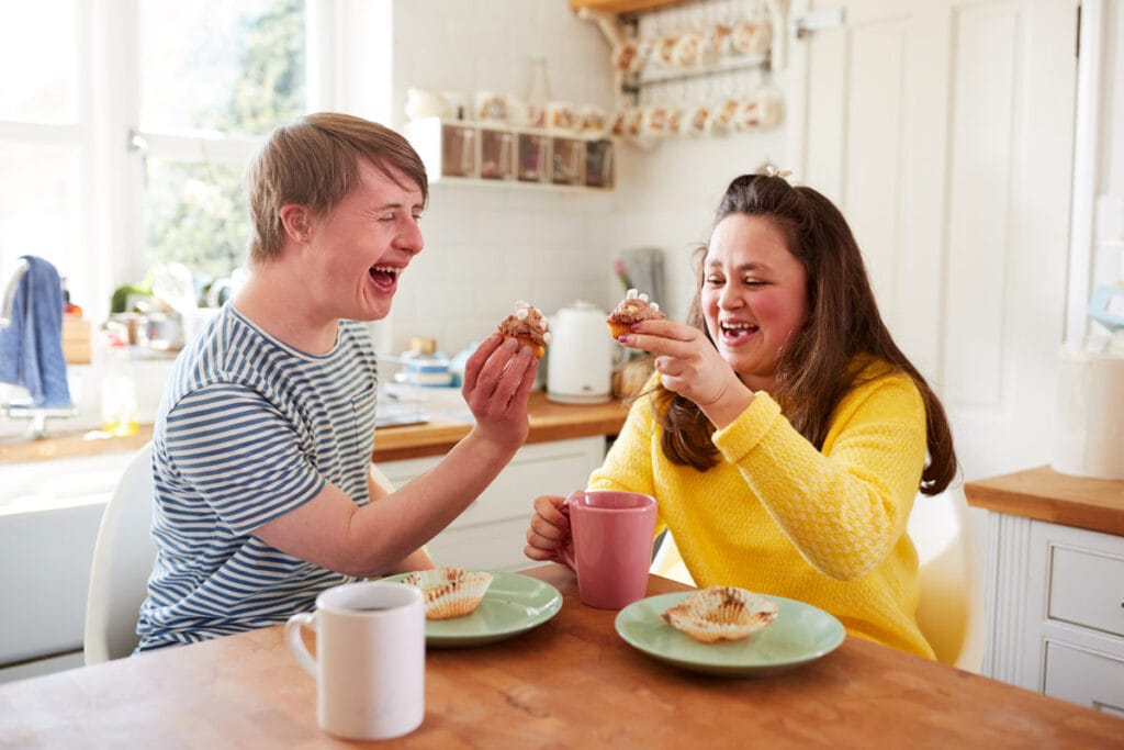 Two people sit together at a table in a kitchen sharing a snack. The man is wearing a striped tshirt and has medium length straight dirty blonde hair. The woman is wearing a bright yellow sweater and has straight brown hair that is down past her shoulders. She has one hand on a pink mug and her other hand is holding up her cupcake like she is about to do a "cheers" with the man and his cupcake.