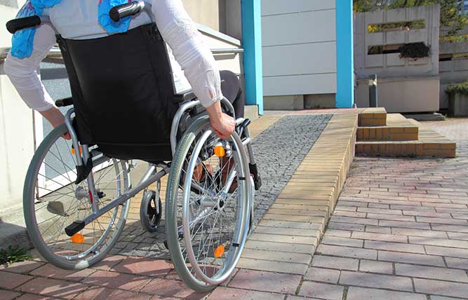 Man in wheelchair proceeding up a ramp to a building entrance.